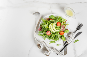 Teller mit Blattsalat, Avocado und Tomaten neben einem Maßband und Besteck auf einem hellen Tisch.