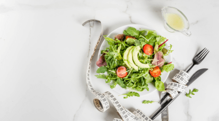 Teller mit Blattsalat, Avocado und Tomaten neben einem Maßband und Besteck auf einem hellen Tisch.