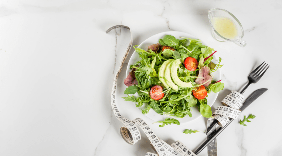 Teller mit Blattsalat, Avocado und Tomaten neben einem Maßband und Besteck auf einem hellen Tisch.
